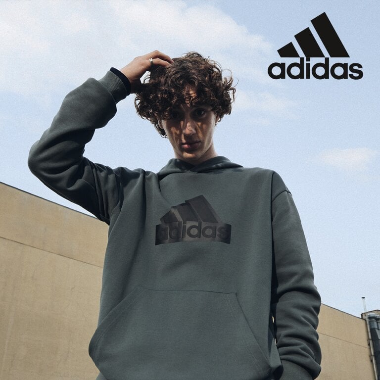 adidas sportswear. Eine Person in einem dunklen adidas Hoodie steht vor einem beigen Gebäude.