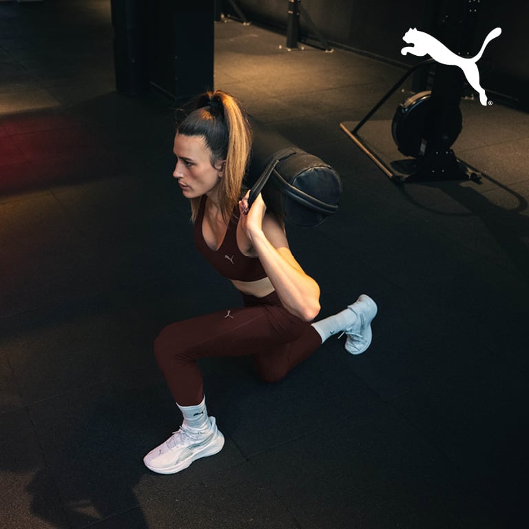 Eine Person macht in einem Fitnessstudio einen Ausfallschritt mit einem Sandsack auf den Schultern. Rechts oben im Bild ist das PUMA‑Logo zu sehen.