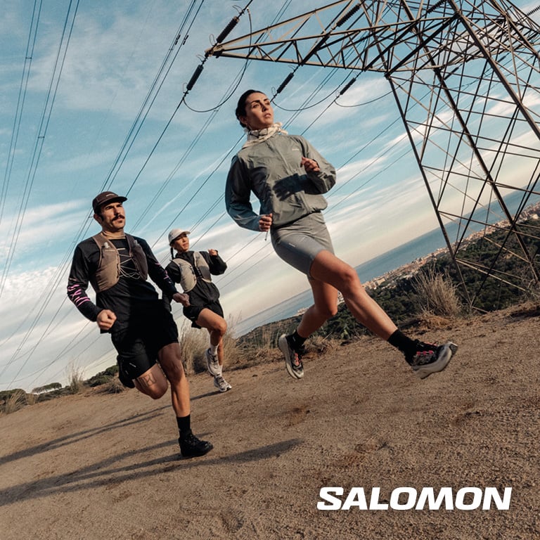 Drei Personen laufen auf einem breiten Schotterweg unter Stromleitungen durch eine hügelige Landschaft. Rechts unten im Bild steht das SALOMON‑Logo.