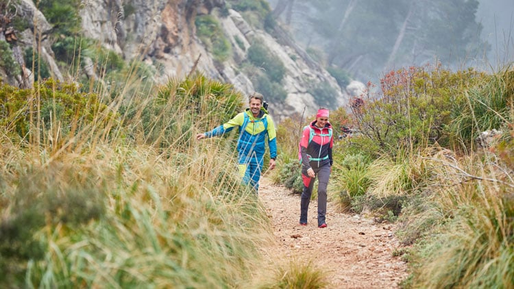 Zwei Wanderer auf einem schmalen Pfad in einer bergigen, bewaldeten Landschaft. Die linke Person trägt eine blaue Jacke mit gelben Akzenten, die rechte eine pinke Jacke. Beide tragen Rucksäcke. Der Weg ist von hohem Gras und Büschen gesäumt, im Hintergrund sind Felsen und Bäume sichtbar