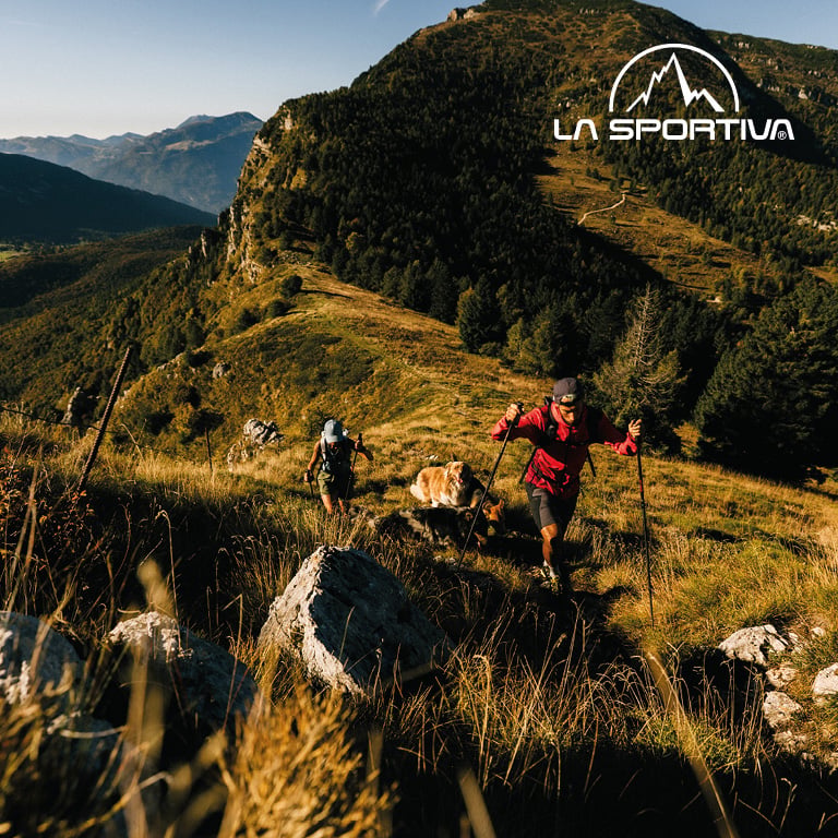 Zwei Personen wandern mit Stöcken einen sonnigen Berghang hinauf, umgeben von Gras und Felsen. Rechts oben im Bild steht das LA SPORTIVA‑Logo.