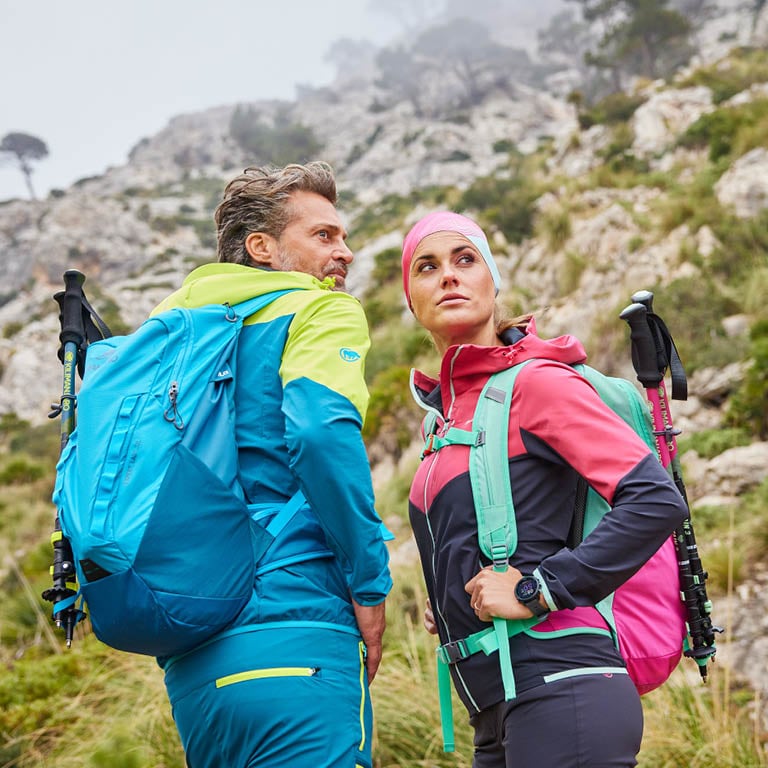 Zwei Personen mit farbigen Outdoor-Rucksäcken und Wanderkleidung stehen in felsiger Berglandschaft.