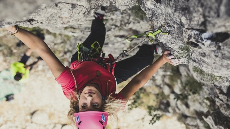 Eine Person beim Felsklettern mit pinkem Helm, rotem Shirt und schwarzer Hose. Sie nutzt Kletterausr&uuml;stung mit Seilen und Karabinern, die an der Felswand befestigt sind. Im Hintergrund ist eine weitere, unscharfe Person zu sehen, vermutlich am Boden oder weiter unten an der Wand.