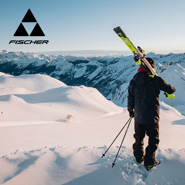 Fischer. Eine Person steht auf einem verschneiten Berghang und hält Skier über der Schulter.