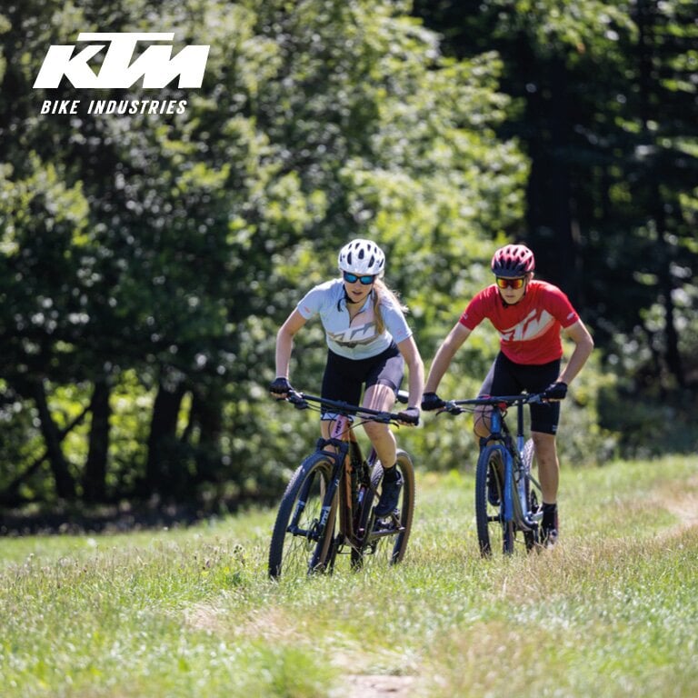 KTM. Zwei Radfahrer fahren mit Mountainbikes auf einem grasbewachsenen Pfad in einem bewaldeten Gebiet.