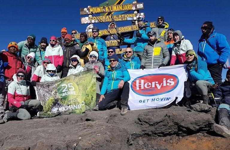 Eine Gruppe Menschen steht am Gipfel des Kilimanjaro. Sie halten Flagge mit Hervis und 25 Jahre Kilimanjaro in die Kamera.