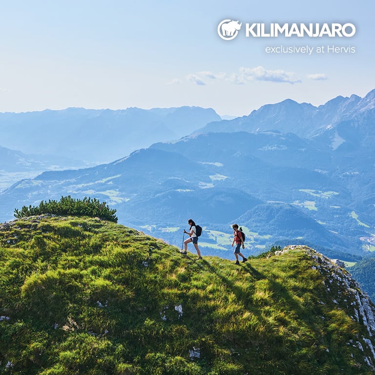Zwei Personen wandern auf einem sonnigen Bergkamm mit weitem Blick über eine hügelige Landschaft. Rechts oben im Bild ist das KILIMANJARO‑Logo mit dem Hinweis „exclusively at Hervis“ zu sehen.