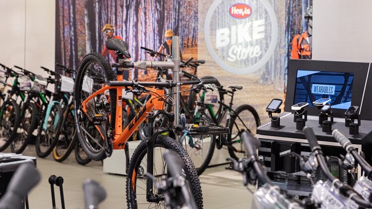 Innenansicht eines ‚Hervis Bike Store‘ mit orangefarbenem Mountainbike auf einem Montageständer im Vordergrund, Wandgrafik mit Radfahrern im Wald im Hintergrund und Verkaufsbereich mit Zubehör auf der rechten Seite.