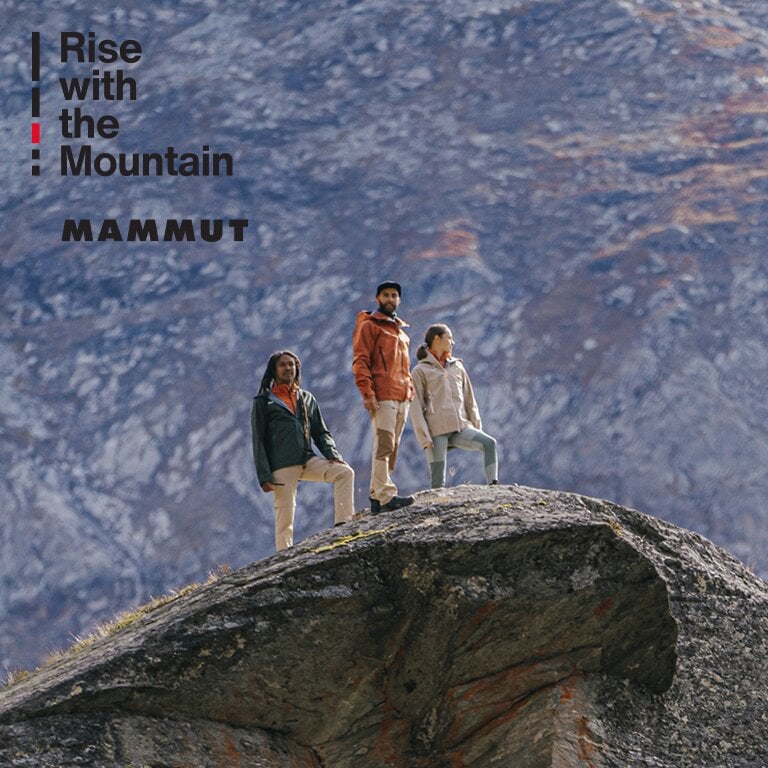 Mammut. Drei Personen stehen auf einem großen Felsen vor einer bergigen Kulisse. Auf der linken Seite des Bildes steht der Text: Rise with the Mountain.