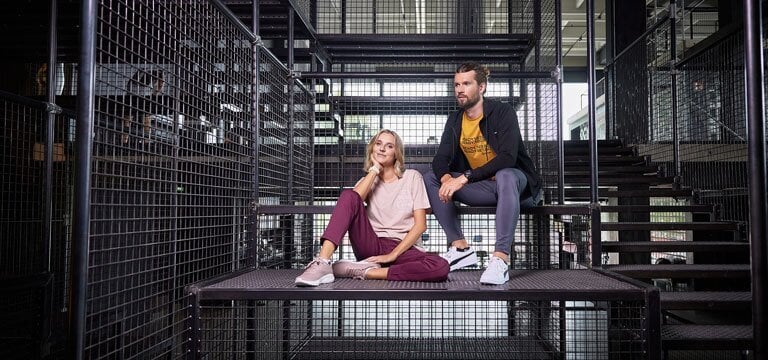 Zwei Personen sitzen auf einer Metallplattform innerhalb einer Struktur aus Metallgittern und Treppen. Eine Person trägt ein rosa Shirt und eine weinrote Hose, die andere eine schwarze Jacke, ein gelbes Shirt und graue Hosen.