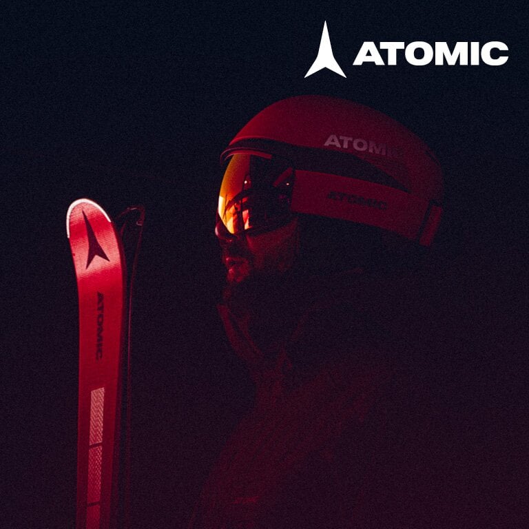 Atomic. Eine Person trägt einen roten Helm und Skibrille und hält einen roten Ski mit der Marke 'ATOMIC'. Der Hintergrund ist dunkel, und das Licht hebt die Person und den Ski hervor.