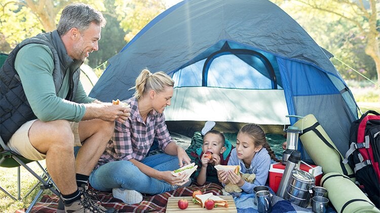 Vier Personen sitzen vor einem Zelt auf einer Decke, umgeben von Lebensmitteln und Campingausrüstung. Die Szene wirkt wie ein gemeinsames Picknick während eines Campingausflugs