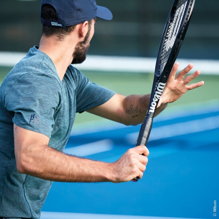 Wilson. Eine Person spielt Tennis auf einem Außenplatz. Sie trägt ein blaues Shirt und eine blaue Kappe und hält einen Wilson-Tennisschläger in der Hand.