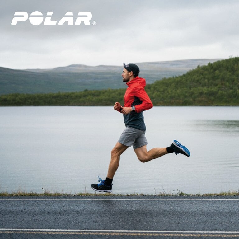 Polar. Eine Person läuft auf einer Straße neben einem See mit Hügeln im Hintergrund. Die Person trägt eine rote Jacke, graue Shorts und blaue Laufschuhe.