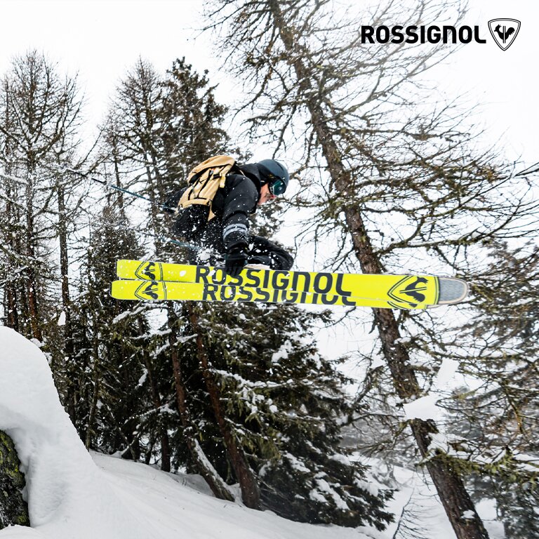 Rossignol. Ein Skifahrer führt einen Sprung in einem verschneiten, bewaldeten Gebiet aus. Die Skier sind gelb und tragen den Markennamen ROSSIGNOL.