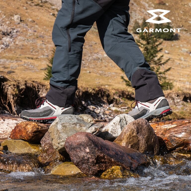 Garmont. Eine Person trägt Wanderschuhe und dunkle Hosen und tritt auf Felsen und Baumstämme in einer natürlichen Umgebung.
