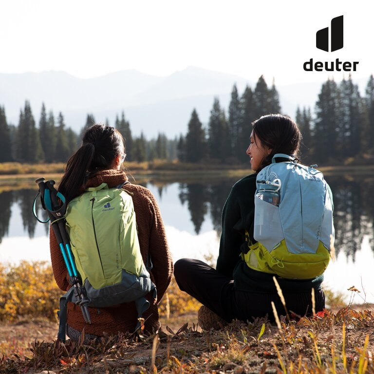 Deuter. Zwei Frauen mit Deuter Rucksäcken sitzen an einem Seeufer.