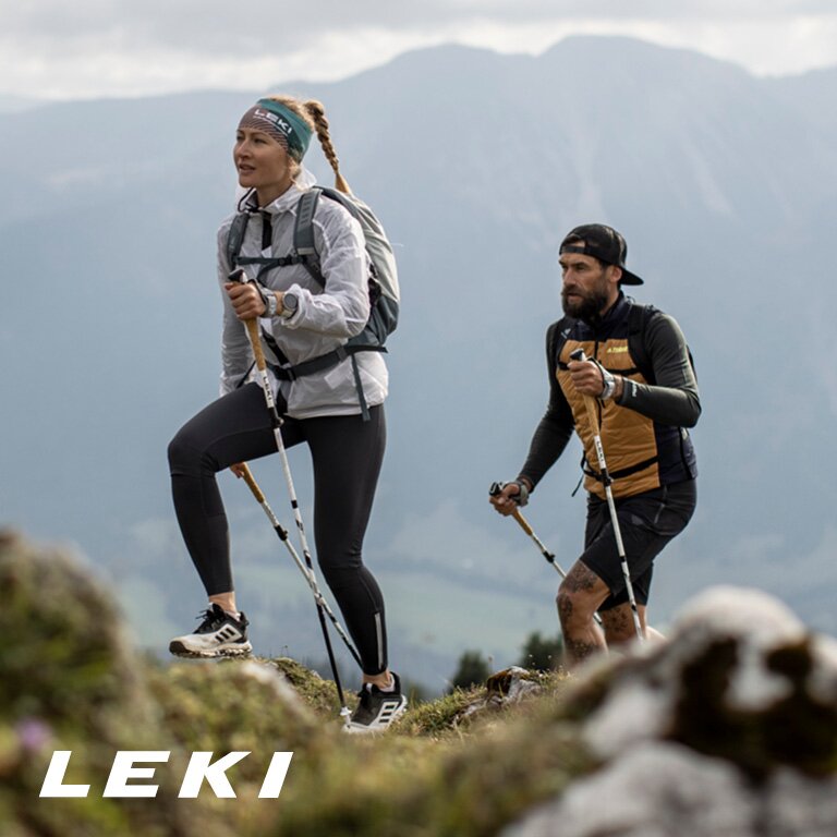 Leki. Zwei Personen wandern in einem bergigen Gebiet. Sie benutzen Trekkingstöcke und tragen Outdoor-Ausrüstung, einschließlich Rucksäcken und Sportkleidung.