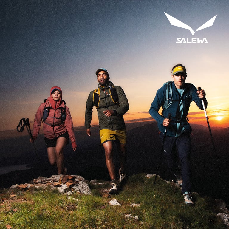 Drei Personen wandern mit Ausrüstung bei Sonnenuntergang über einen felsigen Berggrat. Rechts oben im Bild ist das SALEWA‑Logo zu sehen.