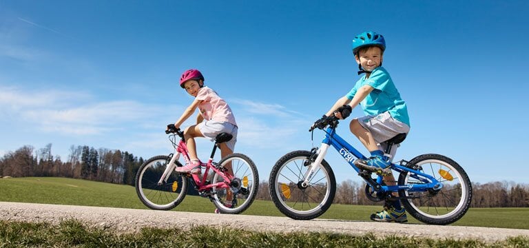 Zwei Kinder fahren auf einem asphaltierten Weg durch ein offenes Feld Fahrrad. Eines fährt ein pinkes Rad mit passendem Helm, das andere ein blaues Rad mit blauem Helm. Beide tragen Shorts und T-Shirts.