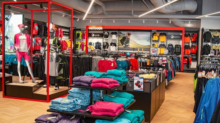 Outdoorberater Innenansicht eines modernen Bekleidungsgeschäfts mit Fokus auf Outdoor- und Sportmode. Bunte Kleidungsstücke wie Jacken, Shirts und Hosen sind ordentlich auf Regalen und Kleiderständern präsentiert. Links steht eine Schaufensterpuppe in pinker Sportkleidung. Der Laden ist hell beleuchtet, mit Holzfußboden und Beschilderungen wie ‚OUTDOOR‘ und ‚MEN‘