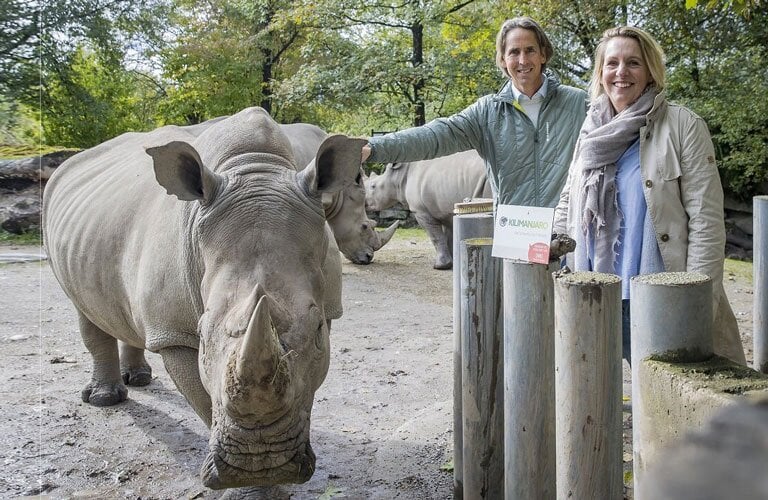 Zwei Personen stehen neben einem Nashorn in einem Zoo. Ein Schild mit dem Schriftzug Kilimanjaro ist zu sehen.