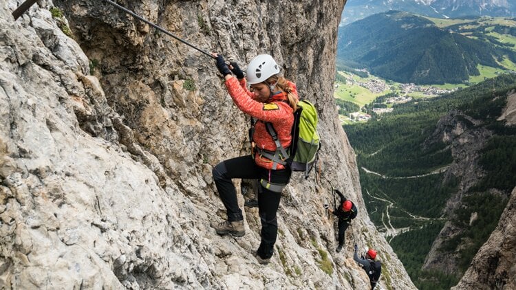 Drei Kletterer an einer steilen Felswand, gesichert mit einem Fixseil. Der vordere Kletterer tr&auml;gt eine rote Jacke und einen gr&uuml;nen Rucksack. Alle sind mit Helmen, Gurten und Rucks&auml;cken ausgestattet. Im Hintergrund erstreckt sich eine weite Landschaft mit gr&uuml;nen T&auml;lern und fernen Bergen unter klarem Himmel.