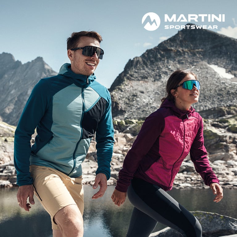 Zwei Personen bewegen sich über felsiges Gelände an einem Bergsee, umgeben von steilen Gipfeln. Rechts oben im Bild steht das MARTINI Sportswear‑Logo.