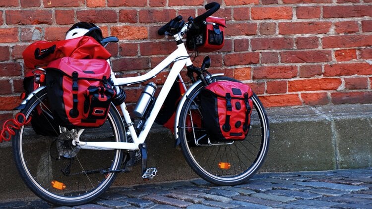 Weißes Fahrrad mit mehreren roten Gepäcktaschen an Vorder- und Hinterrad sowie einer kleinen Lenkertasche lehnt an einer Backsteinmauer; ein Fahrradhelm liegt auf einer der hinteren Taschen.