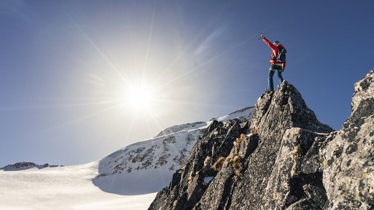 Eine Person steht auf einem felsigen Berggipfel, hebt ein Eispickel in die Luft und trägt eine rote Jacke sowie einen Rucksack. Die Sonne scheint am klaren blauen Himmel und beleuchtet die schneebedeckte Landschaft und das felsige Gelände.