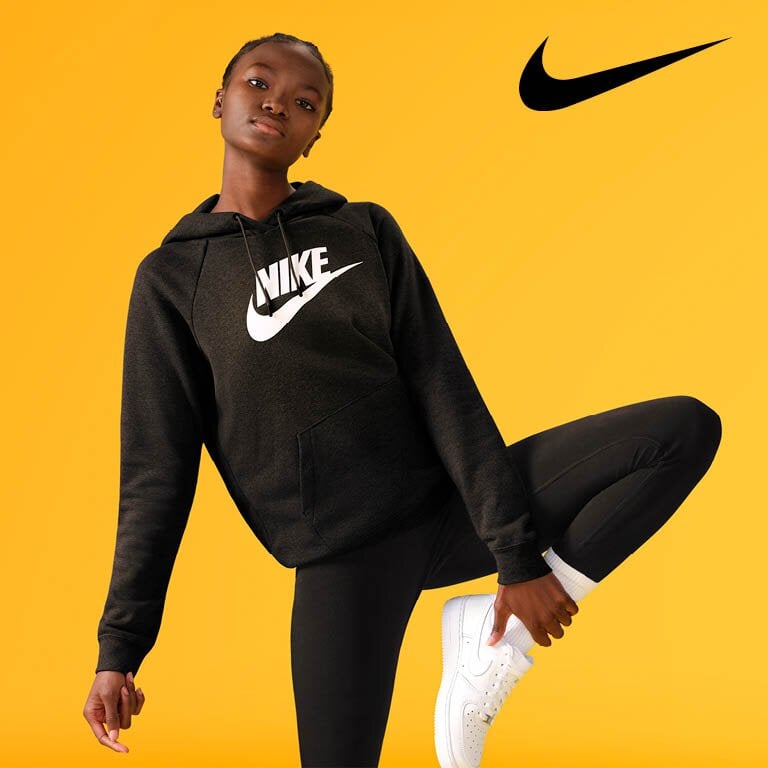 Nike. Eine Person trägt einen schwarzen Nike-Hoodie und schwarze Hosen und steht auf einem Bein, während sie das andere Bein mit einer Hand hochhält.