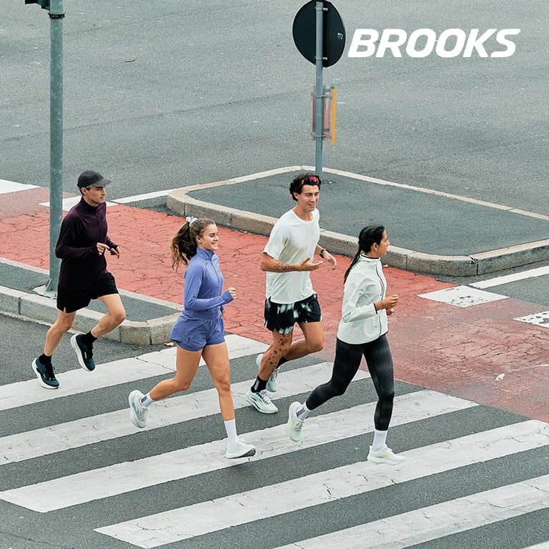 Vier Personen laufen gemeinsam über einen breiten Zebrastreifen in einer urbanen Umgebung. Rechts oben im Bild steht das BROOKS‑Logo.