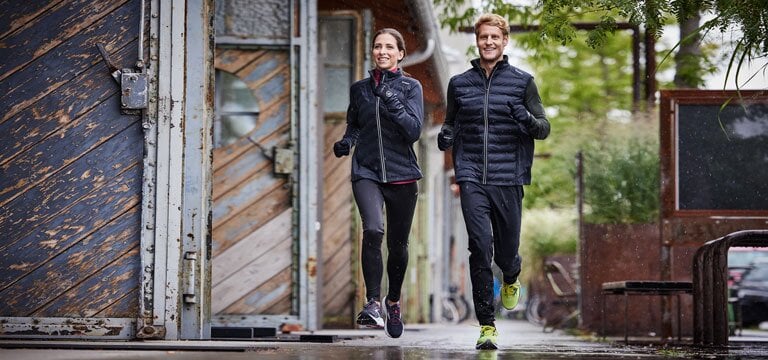 Zwei Personen joggen auf nassem Pflaster in einer städtischen Umgebung. Beide tragen dunkle Sportkleidung. 