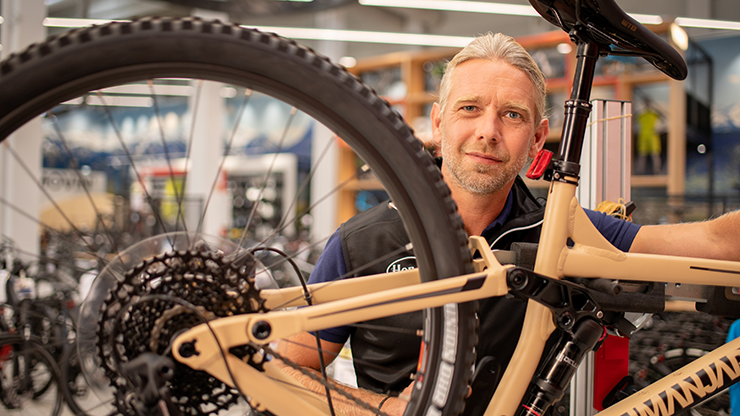 Eine Person arbeitet an einem beigefarbenen Fahrrad in einer Fahrradwerkstatt oder einem Reparaturbereich. Das Gesicht der Person ist unscharf, und der Fokus liegt auf dem Hinterrad und dem Getriebemechanismus des Fahrrads. Im Hintergrund sind weitere Fahrräder und Ausrüstungen zu sehen.
