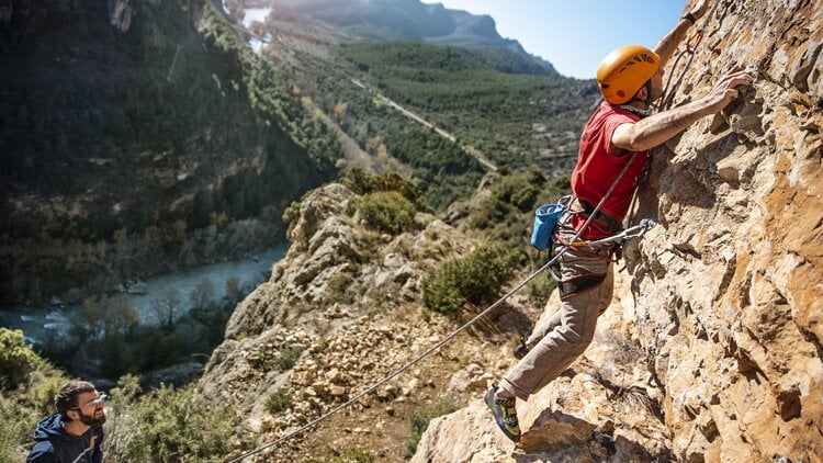 Zwei Personen beim Felsklettern. Eine Person mit gelbem Helm, rotem Shirt und Klettergurt steigt eine Felswand hinauf, während die andere unten steht und möglicherweise sichert. Im Hintergrund ist ein Tal mit Fluss, dichter Vegetation und fernen Bergen unter klarem Himmel zu sehen.