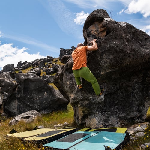 Eine Person beim Bouldern an einer großen Felsformation im Freien. Sie trägt ein orangefarbenes Shirt und grüne Hose. Unter ihr liegen Crashpads zur Absicherung. Im Hintergrund sind weitere Felsen und ein teils bewölkter Himmel zu sehen.