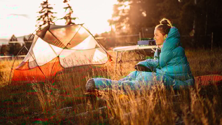 Person sitzt in einem blauen Schlafsack auf einer Wiese im Freien. In der Nähe steht ein orange-weißes Zelt. Die Szene spielt bei Sonnenauf- oder -untergang, mit warmem Licht, Bäumen im Hintergrund und Campingausrüstung auf einem Tisch.