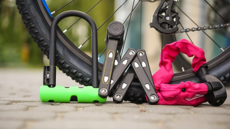 Nahaufnahme eines Fahrradlaufrads mit drei verschiedenen Fahrradschlössern im Vordergrund: ein grünes Bügelschloss, ein schwarzes Faltschloss und ein pinkes Kettenschloss.