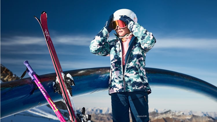 Kind in Winterkleidung mit Tarnjacke, dunkler Hose und weißem Helm steht in einer verschneiten Berglandschaft und richtet den Helm mit beiden Händen. Neben ihr lehnen zwei Paar Skier an einer Metallstruktur. Im Hintergrund sind schneebedeckte Berge unter klarem, blauem Himmel zu sehen.