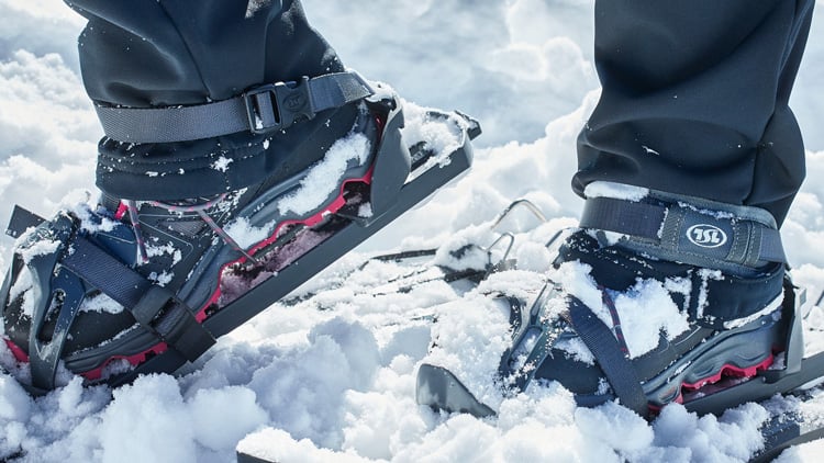 Nahaufnahme von zwei Füßen mit Schneeschuhen, die auf Schnee stehen. Die Schneeschuhe haben schwarze und rote Riemen und sind teilweise mit Schnee bedeckt. Die Person trägt schwarze Hosen