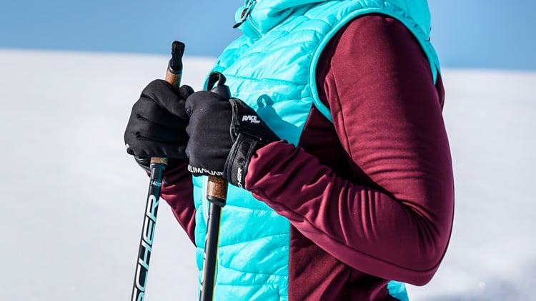 Eine Person in Winterkleidung steht in einer verschneiten Landschaft. Sie trägt eine türkisfarbene Steppweste über einem weinroten Langarmshirt und hält Skistöcke mit schwarzen Handschuhen