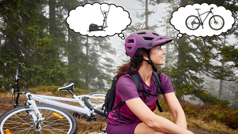 Eine Frau in lila Radkleidung und Helm sitzt im Wald auf dem Boden, neben ihr liegt ein weißes Fahrrad. Über dem Kopf befinden sich zwei Gedankenblasen: eine mit einem Crosstrainer, die andere mit einem Fahrrad.