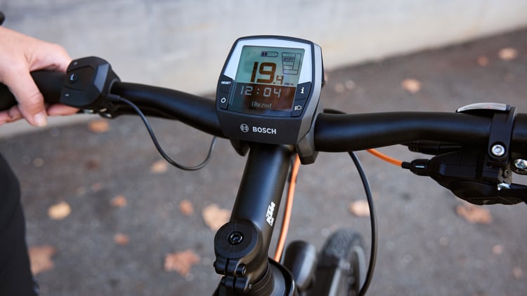Nahaufnahme der Fahrradlenker mit einem Bosch-Display, das Geschwindigkeit (19,4 km/h), Uhrzeit (12:04) und weitere Fahrdaten anzeigt; links Bedieneinheit mit Tasten, rechts Bremshebel und Schalthebel.