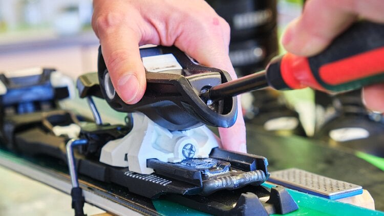 Eine Nahaufnahme einer Person, die mit einem Schraubendreher die Bindung an einem Ski einstellt. Eine Hand der Person hält die Bindung, während die andere Hand den Schraubendreher benutzt. Der Ski und die Bindung sind deutlich sichtbar, während einige Hintergrundelemente leicht unscharf sind.