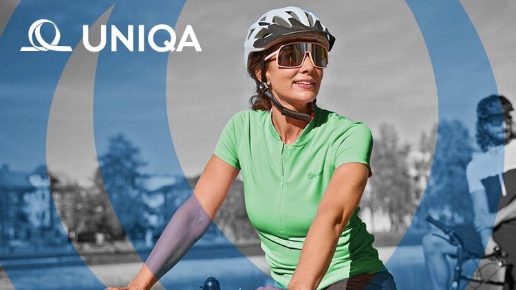 Eine Person trägt ein grünes Shirt und einen weißen Helm und fährt Fahrrad. Im Hintergrund ist ein weiterer Radfahrer und einige Bäume zu sehen. Das Logo 'UNIQA' ist oben links im Bild gut sichtbar.