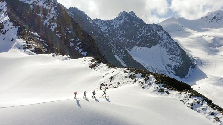 Eine Gruppe von vier Personen besteigt einen schneebedeckten Berg. Die Personen gehen in einer Reihe, und ihre Schatten sind auf dem Schnee sichtbar. Im Hintergrund sind zerklüftete, felsige Berge mit Schneefeldern und Eis unter einem teilweise bewölkten Himmel zu sehen.