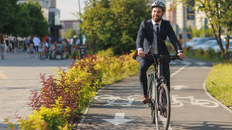 Person im Anzug und mit Helm fährt auf einem Fahrrad entlang eines markierten Radwegs mit weißen Fahrradsymbolen und Richtungspfeilen. Am Rand des Weges befinden sich Büsche, im Hintergrund sind Fußgänger zu sehen.