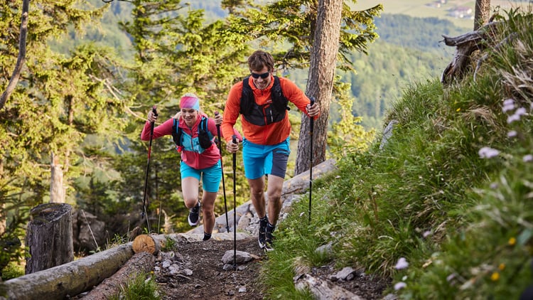 Zwei Wandernde mit Trekkingstöcken und Rucksäcken steigen einen felsigen Waldpfad hinauf; vorne eine Person in orangefarbenem Shirt und blauer Shorts, dahinter eine in pinkem Shirt und blauer Shorts, umgeben von grünen Bäumen und Berglandschaft