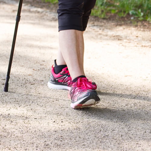Nahaufnahme einer Person beim Gehen mit Walking-Stöcken auf einem Kiesweg. Sie trägt schwarze Caprihosen und pink-schwarze Sportschuhe