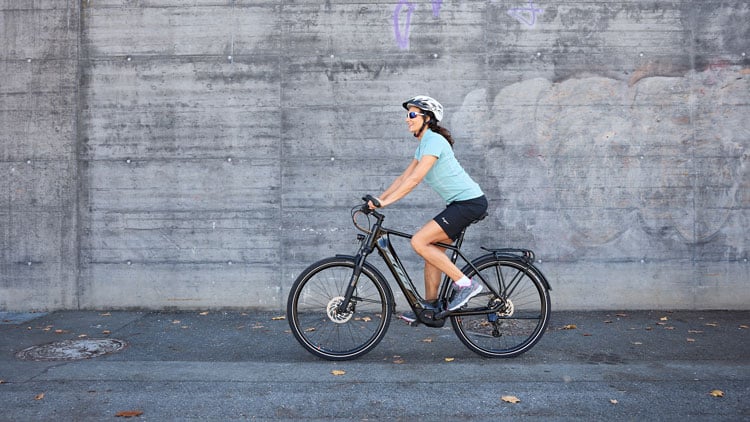 Person fährt mit einem E-Bike auf einem gepflasterten Untergrund; im Hintergrund eine Betonwand. Die Person trägt einen Helm, ein hellblaues Radshirt, schwarze Radhoshorts, sowie weiße Socken und Sneakers.
